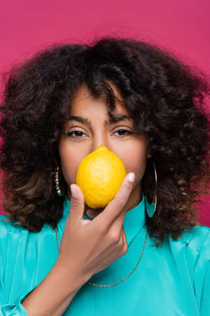 african american woman with wavy hair obscuring face with lemon isolated on pink