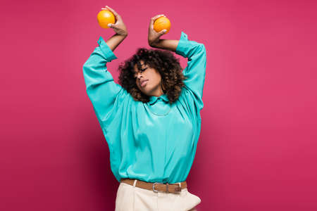 african american woman in turquoise blouse posing with oranges in raised hands  isolated on pinkの写真素材