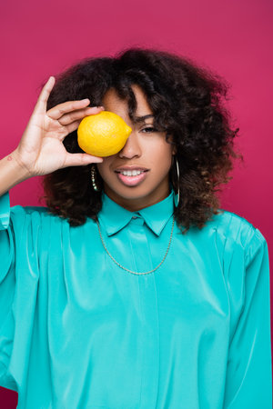 african american woman in blue blouse covering eye with lemon isolated on pinkの写真素材