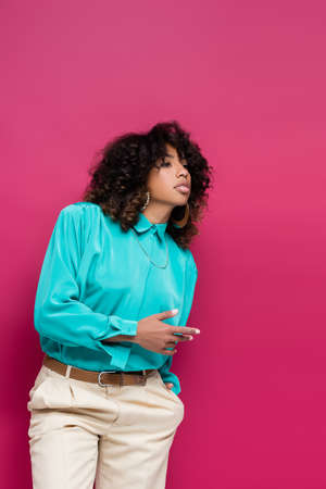 african american woman in turquoise blouse standing with hand in pocket and looking away isolated on pinkの写真素材