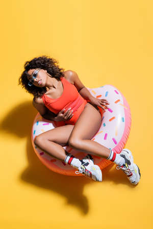 top view of african american woman in trendy sunglasses and beachwear sitting on swim ring on yellow backgroundの写真素材