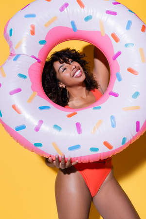cheerful african american woman with curly hair holding inflatable ring on yellow background