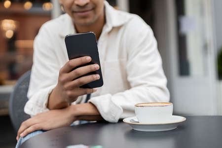 cropped view of blurred african american man texting on smartphone near cup of cappuccino on table...