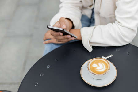 partial view of african american man texting on smartphone near cup of cappuccino on table