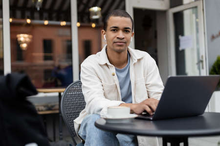 african american freelancer in wireless earphones sitting near cup of coffee and laptop on summer...