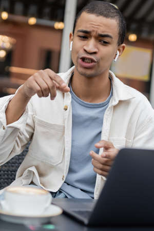 african american freelancer in wireless earphones gesturing during video call on summer terrace