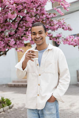 young african american man holding takeaway drink near pink cherry tree on street