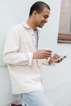 happy african american man holding paper cup and smartphone near building on street in europeの写真素材