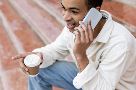 african american man holding takeaway beverage and talking on smartphone while sitting on stairs in...