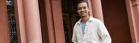 joyful african american man in shirt jacket standing near building, banner