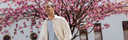 positive african american man in shirt jacket walking near cherry tree, banner