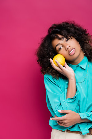 african american woman with wavy hair holding ripe orange isolated on pink