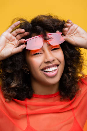joyful african american woman holding stylish sunglasses and looking at camera isolated on yellow