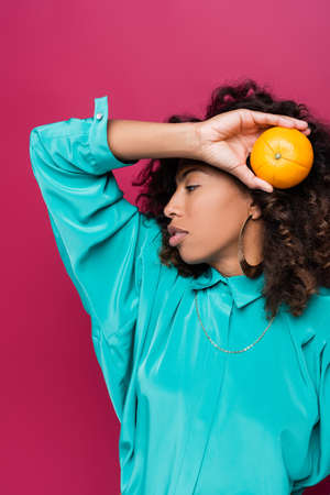 curly african american woman posing with ripe orange isolated on pinkの写真素材