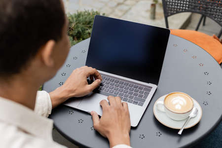 african american freelancer using laptop near cup of latte with foam on table in cafe terrace