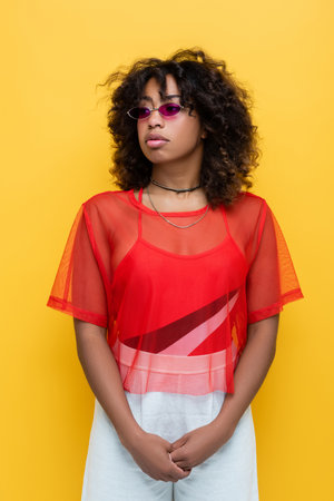 curly african american woman in trendy sunglasses and summer outfit isolated on yellow