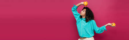 stylish african american woman in blue blouse posing with oranges isolated on pink, banner