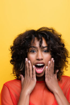amazed african american woman touching face and looking at camera isolated on yellow