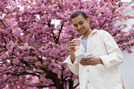 low angle view of happy african american man holding coffee to go and using smartphone near pink cherry treeの写真素材