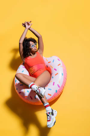 top view of african american woman in swimsuit and stylish sunglasses sitting on swim ring on yellow backgroundの写真素材