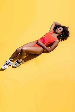 full length of african american woman in red swimsuit, sunglasses and sneakers on yellow background,...