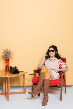 Smiling asian woman in sunglasses and vintage clothes talking on telephone while sitting on armchair on orange backgroundの写真素材
