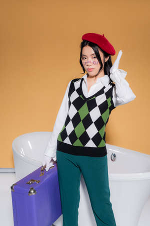 Stylish asian model in beret holding suitcase near bathtub on orange background