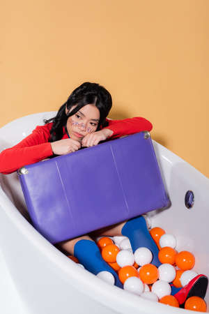 Pretty asian model with glitter on face holding retro suitcase in bathtub with balls on orange...