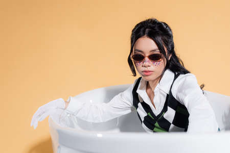 Fashionable asian woman in sunglasses and glitter on face looking at camera in bathtub on orange...