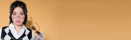 Stylish asian woman holding sunglasses and looking at camera on orange background, bannerの写真素材