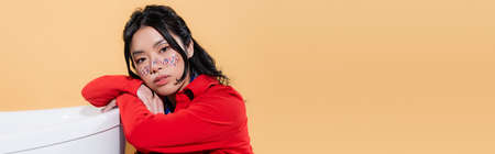 Asian model with glitter on face looking at camera near bathtub isolated on orange, bannerの写真素材