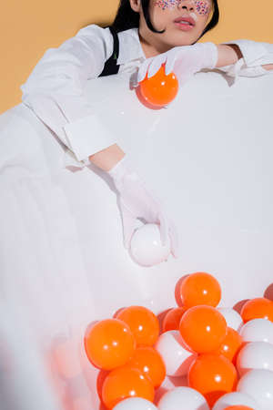 Cropped view of young stylish woman in gloves holding balls near bathtub on orange backgroundの写真素材
