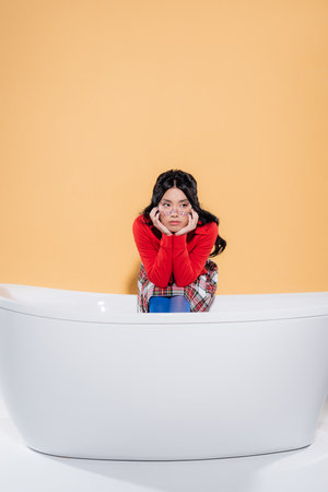 Stylish asian model sitting on bathtub on orange background