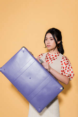 Young asian woman in retro clothes holding suitcase on orange backgroundの写真素材