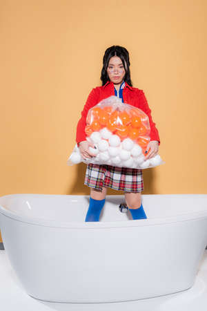 Stylish asian woman holding plastic bag with balls and looking at camera in bathtub on orange backgroundの写真素材