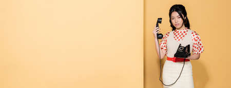 Young asian model in retro clothes holding telephone on orange background, bannerの写真素材