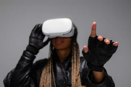 Blurred african american woman in virtual reality headset and gloves pointing with finger isolated on greyの写真素材