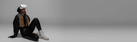 Full length of african american woman in vr headset and leather jacket sitting on grey background,...