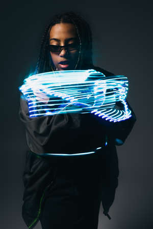 Stylish african american woman in smart glasses looking at neon lights on grey backgroundの写真素材