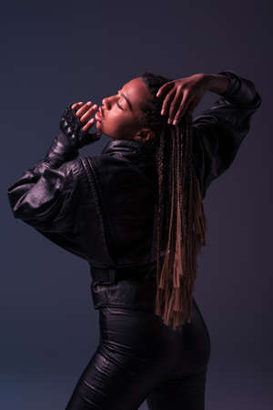 African american woman in glove and leather jacket touching pigtails isolated on dark grey