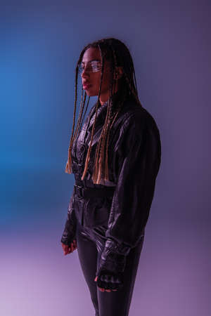 Stylish african american woman in leather jacket and smart glasses standing on blue and purple...