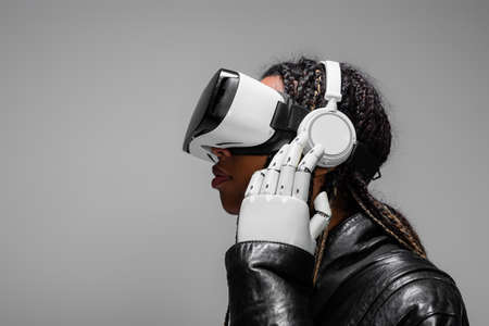 Side view of african american woman with robotic hand and vr headset listening music in headphones...