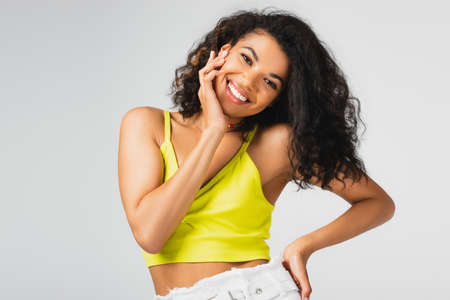 happy african american woman in yellow crop top posing with hand on hip isolated on grey