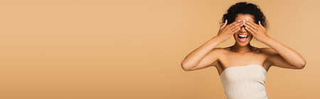 excited african american woman with bare shoulders covering eyes with hands isolated on beige, bannerの写真素材