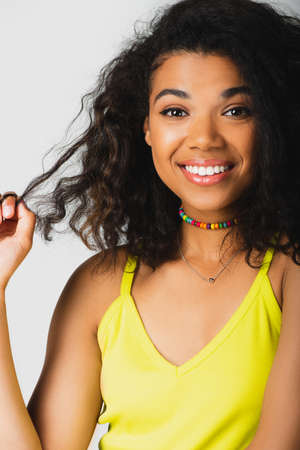 cheerful african american woman pulling curly hair and smiling isolated on grey