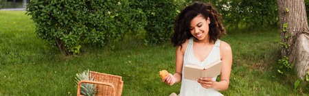 happy african american woman in white dress holding peach and reading book during picnic, bannerの写真素材