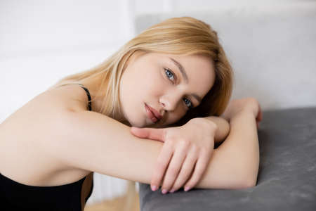 Portrait of blonde woman looking at camera near blurred couchの写真素材