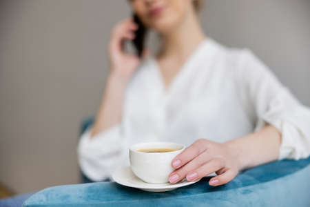Cropped view of blurred woman with smartphone holding cup of coffee on armchairの写真素材