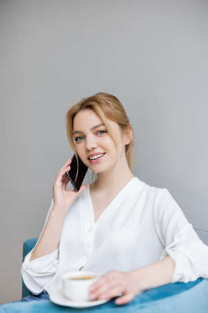 Pretty blonde woman talking on smartphone near blurred coffee cup at home