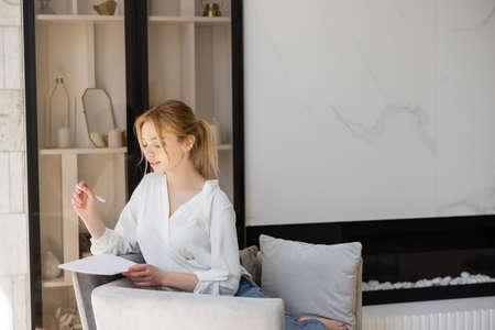 Young woman in white blouse holding notebook and pen on armchair at home